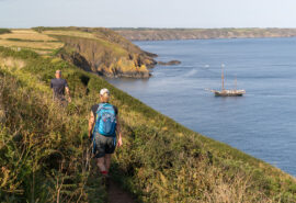 Bessie Ellen guests walking along the Cornish Coast with tall ship anchored, Cornwall,