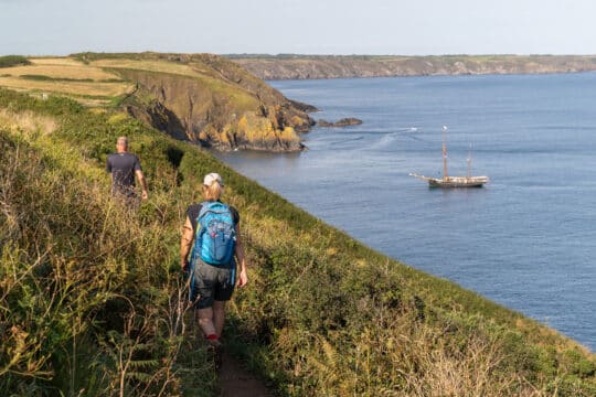 Bessie Ellen guests walking along the Cornish Coast with tall ship anchored, Cornwall,