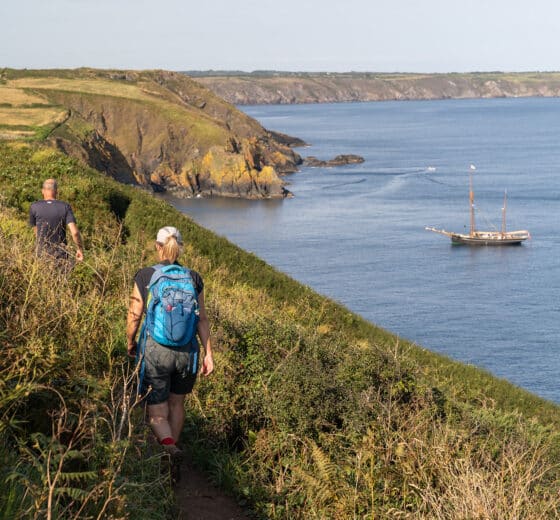 Bessie Ellen guests walking along the Cornish Coast with tall ship anchored, Cornwall,